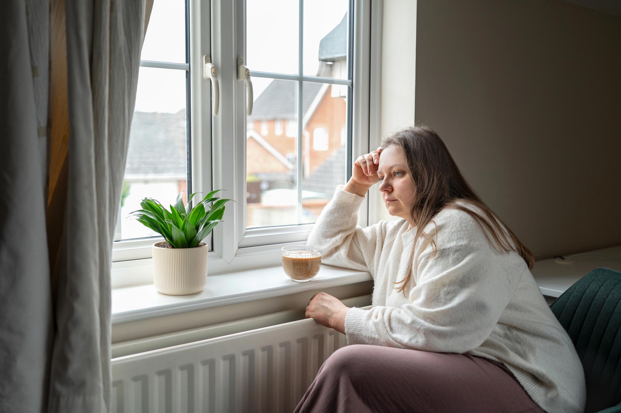 Woman-in-her-40s-standing-in-kitchen-looking-at-her-reflection-in-a-window,-thoughtful-expression-—-perimenopause-weight-gain-and-hormone-imbalance